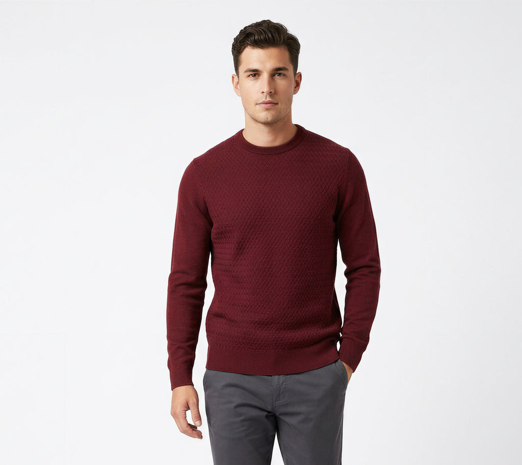 A man modeling a burgundy textured knit crewneck sweater, paired with dark gray trousers and white sneakers, against a plain white studio background.