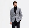 A man modeling a light gray textured blazer over a black turtleneck sweater, paired with black trousers and black leather shoes, against a plain white studio background.