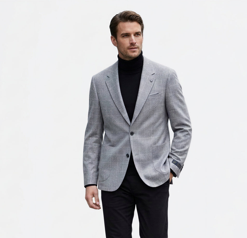 A man modeling a light gray textured blazer over a black turtleneck sweater, paired with black trousers and black leather shoes, against a plain white studio background.