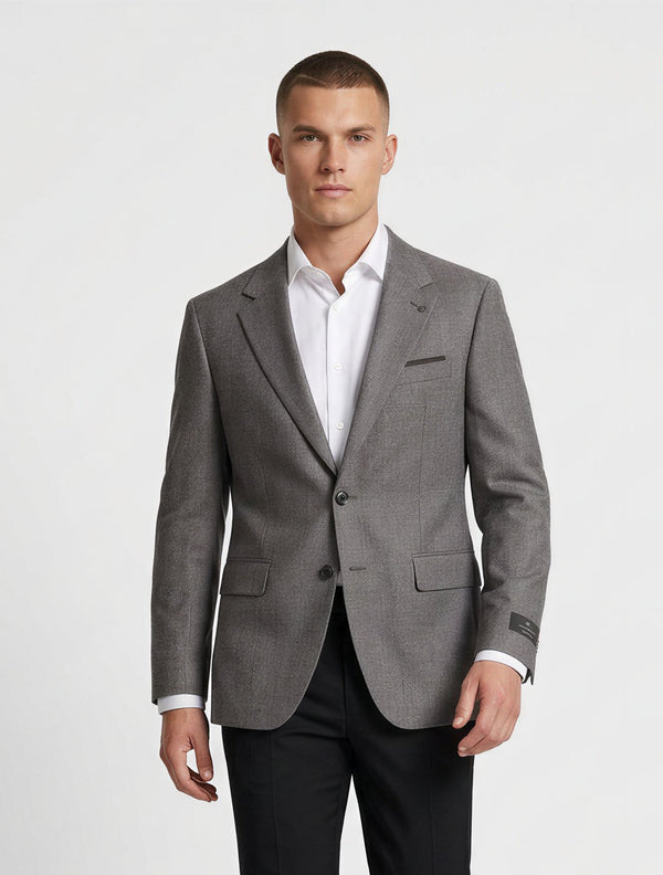 A man modeling a gray two-button suit jacket with black trousers, a white dress shirt, and black dress shoes, standing against a light gray studio background.