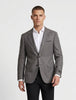 A man modeling a gray two-button suit jacket with black trousers, a white dress shirt, and black dress shoes, standing against a light gray studio background.